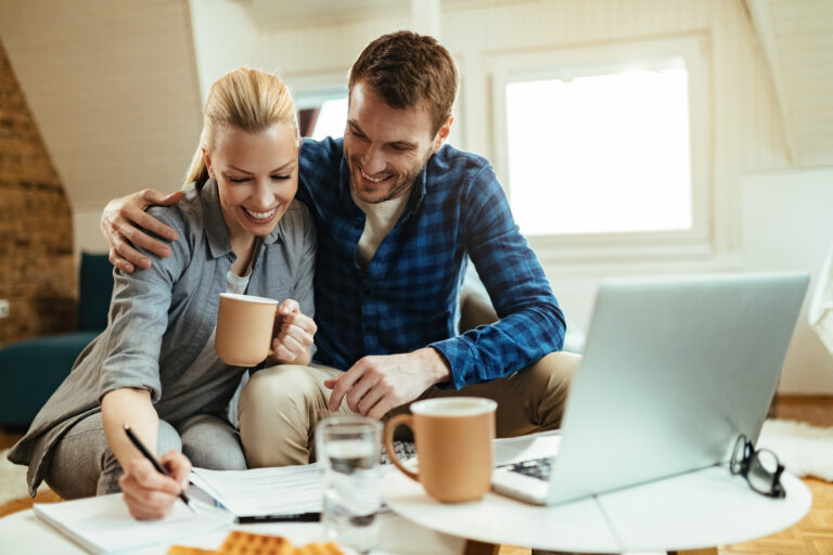 Construção patrimonial: casal sorridente revisando planejamento financeiro pessoal com anotações e laptop em casa.