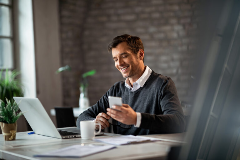 Homem utilizando smartphone ao lado de um notebook, representando o acompanhamento de investimento com liquidez e o planejamento financeiro.